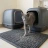 Un elegante gato gris de raza Russian Blue sale de un moderno arenero cubierto y pisa una alfombra atrapa-arena gris texturizada y especializada. La alfombra captura visiblemente pequeños gránulos de arena sueltos de las patas del gato. El piso de baldosas de cerámica circundante en una cocina de Lima está perfectamente limpio.