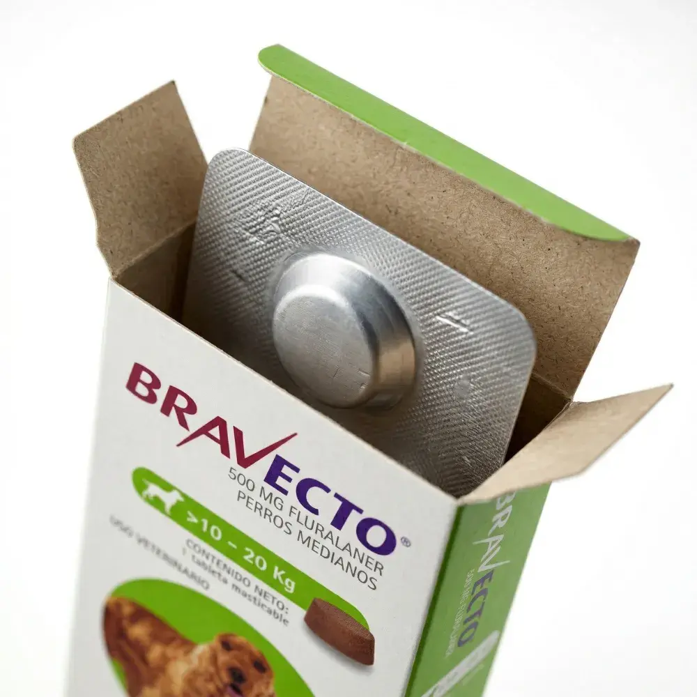 Una fotografía macro de primer plano que muestra una caja abierta de Bravecto para perros medianos (10-20 kg). Se ve el interior de la caja con un blíster de aluminio plateado original y distintivamente grueso parcialmente visible, que contiene una pastilla masticable sellada. Los detalles del empaque exterior son nítidos sobre un fondo blanco y limpio.