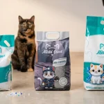 Tres empaques de arena para gatos Mishi Sand de 4kg, 5kg y 10kg en un estudio profesional con un gato sentado al fondo.