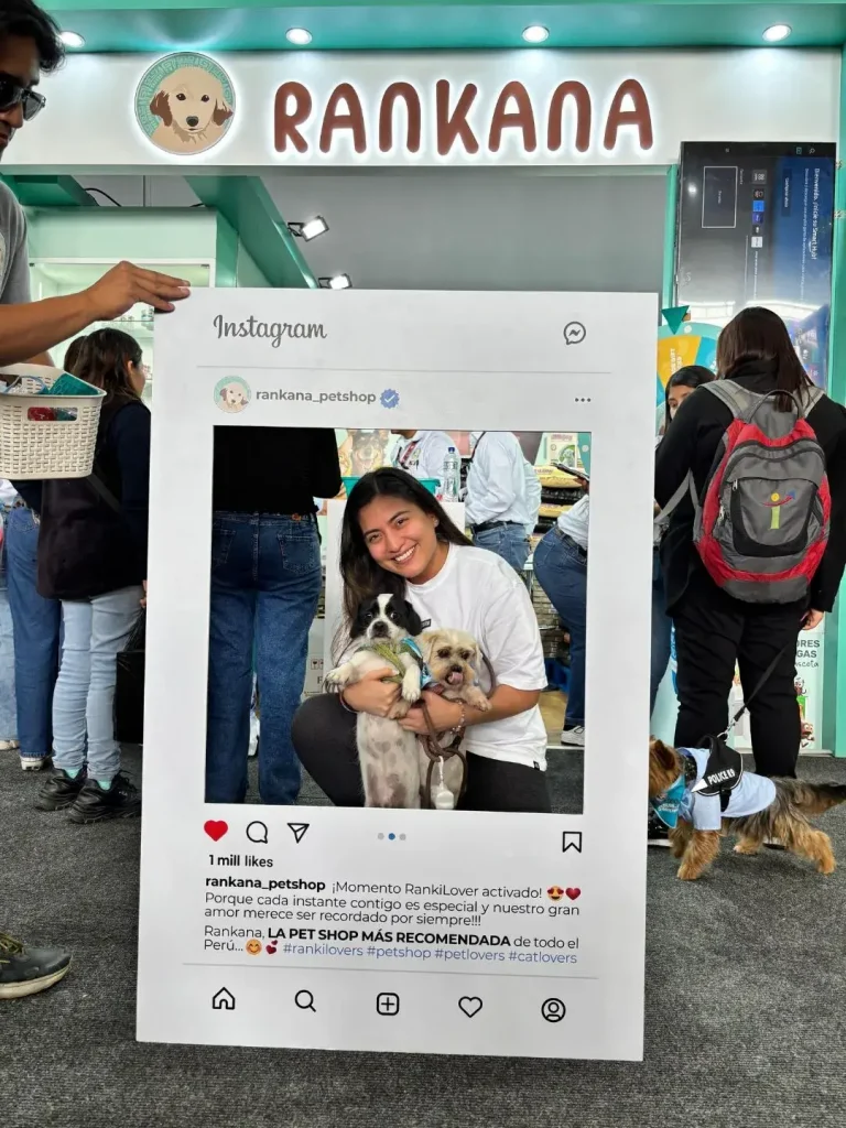 Joven sonriente sosteniendo a dos perritos pequeños en el marco de Instagram de Rankana Pet Shop durante una feria de mascotas en Perú.